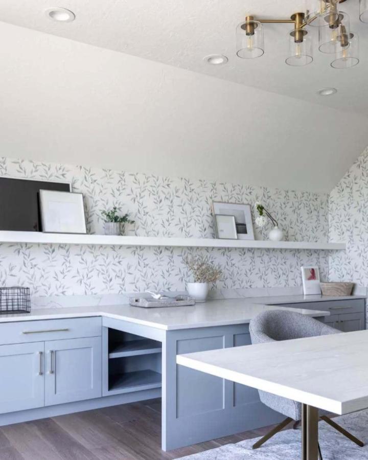 Gray craft room cabinets with botanical wallpaper from @raineyhomesutah. White countertops and floating shelves complete the organized workspace.