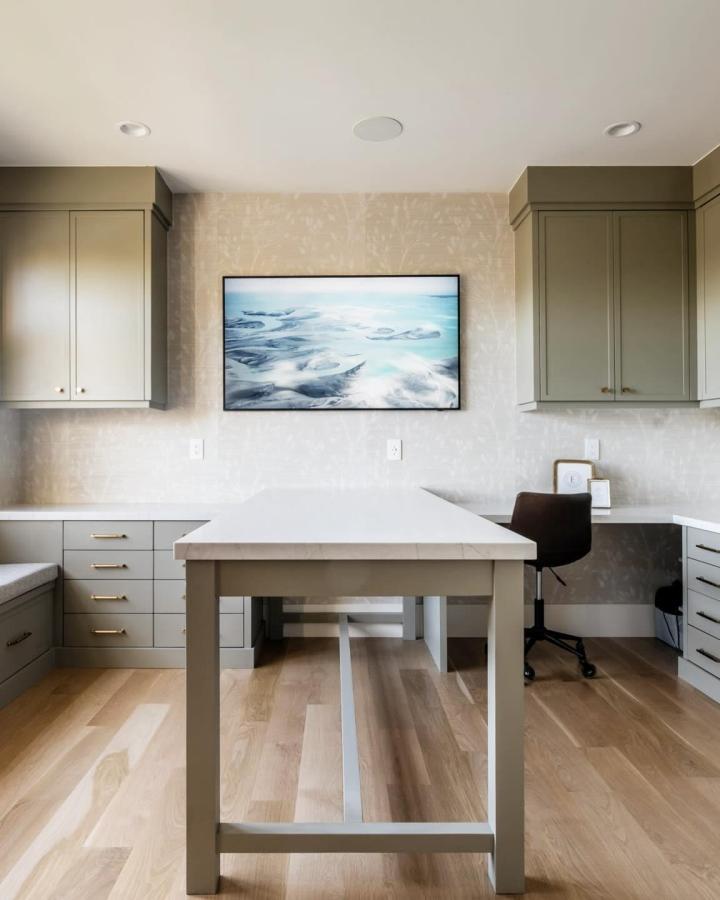 Sage green craft room from @mahoganydesignstudios. Central work table with extensive cabinet storage and corner desk area.