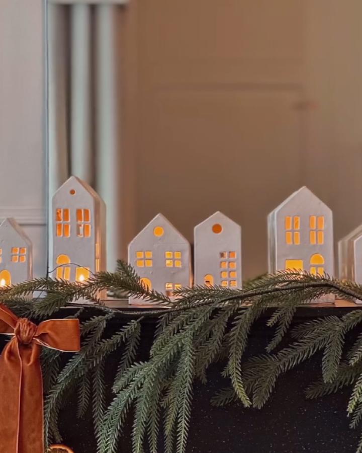 White paper house luminaries from @jellyandjonny. Glowing windows nestle among evergreen branches and ribbon.
