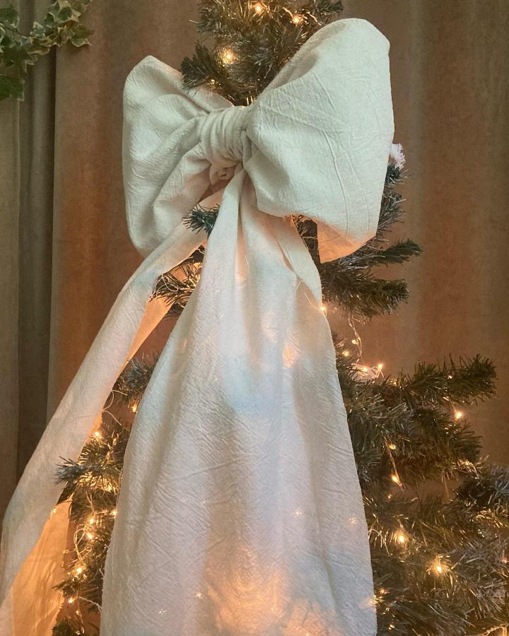 Cream textured fabric bow topper from @paulafellingerinteriores with warm lights and greenery beneath.