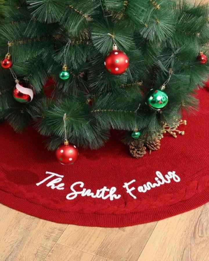 Personalized red knit tree skirt from Zakkaparts. White embroidered family name displayed beneath festive tree.