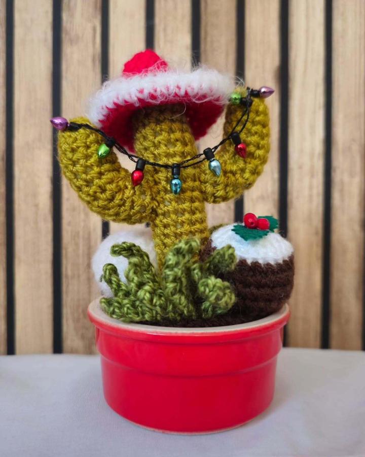 Festive mustard cactus with Santa hat displaying crochet christmas decorations ideas from @thecrochetcactus with string lights and pudding.