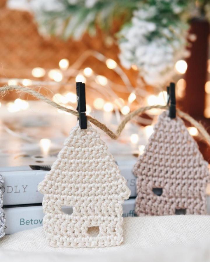 Cream crocheted house ornaments from @lilleliis_official displaying crochet christmas decorations ideas with warm bokeh lights behind.