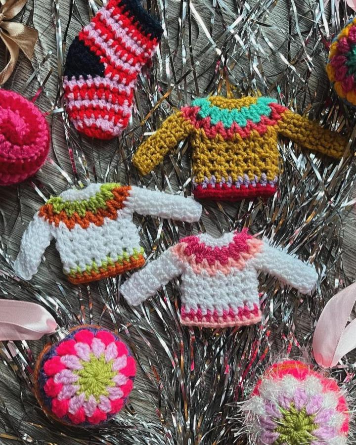 Colorful miniature crocheted sweater ornaments from @foreststitchcraft displayed on silver tinsel garland with festive details.