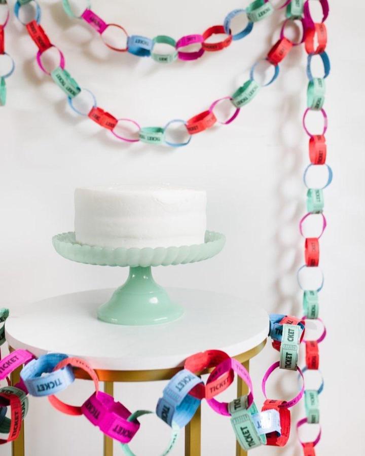 Colorful ticket chain garland from @prettylifegirls draped around white cake on mint stand.