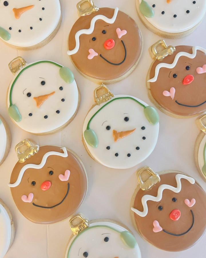 Round decorated cookie ornaments from @bakedgainesville featuring cheerful snowman and gingerbread characters with gold hangers.