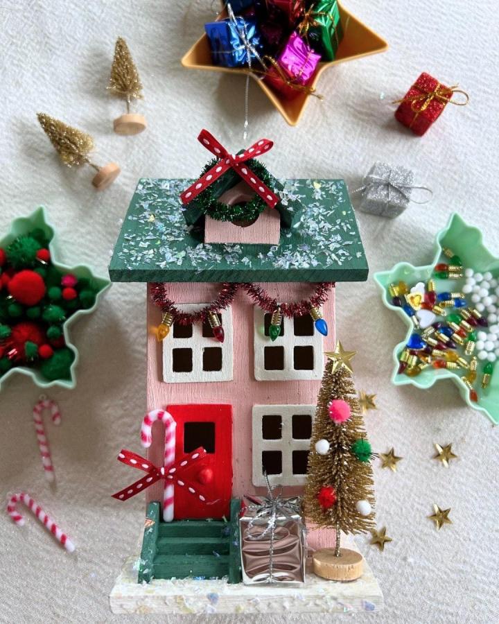 Pink miniature holiday house from Maker Social with green roof and festive embellishments displayed on white surface.