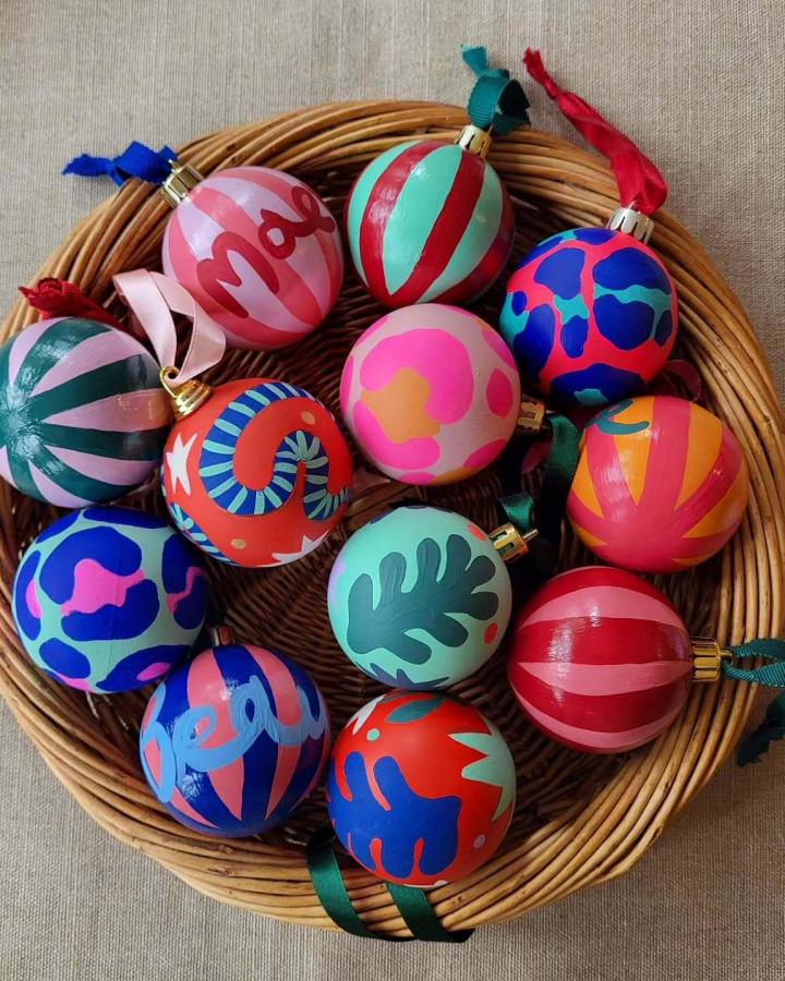 Colorful hand-painted ornaments from @dottie_and_cloud in wicker basket with bold patterns and ribbon hangers