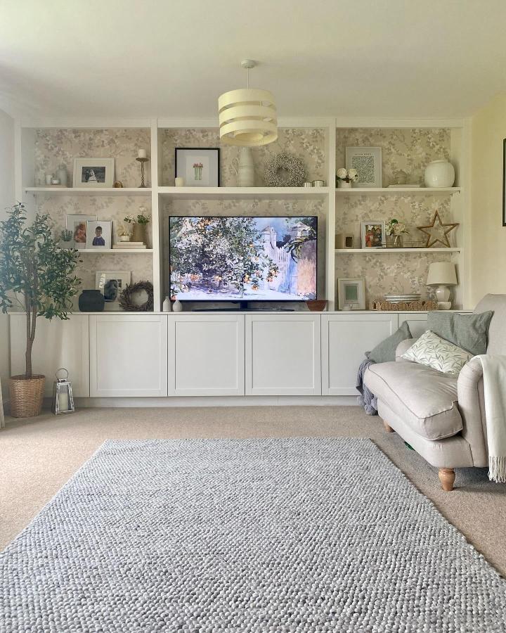 White built-in media unit with floral wallpaper backing from @townhouse_on_the_green. Shelves styled with decor and photos.