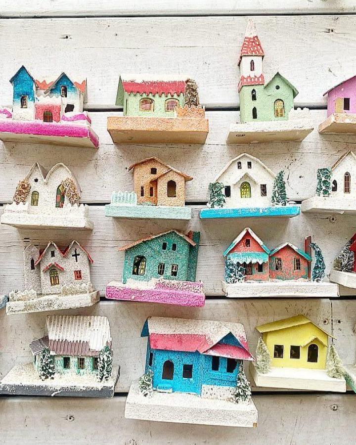 Colorful putz houses from @the_purple_ottoman arranged on white shelves with glittery snow bases and pastel roofs