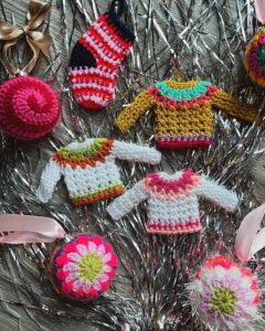 Colorful crocheted sweater ornaments from @foreststitchcraft displayed on silver tinsel garland with festive details.