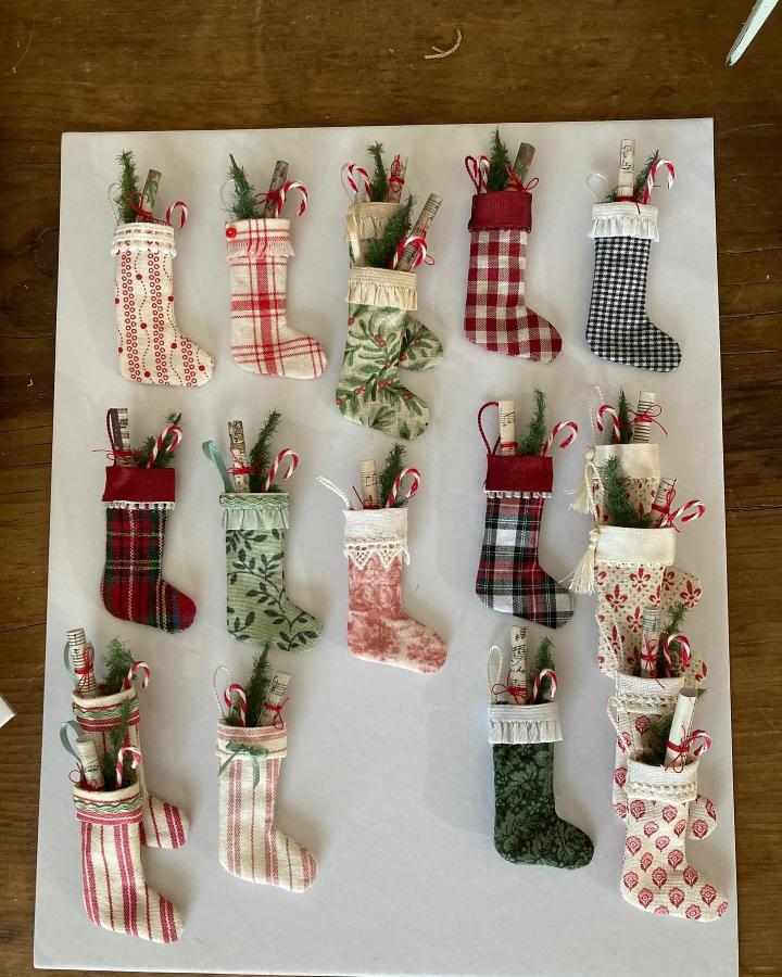 Patterned fabric stockings from @echminiatures filled with candy canes and greenery arranged on white board.