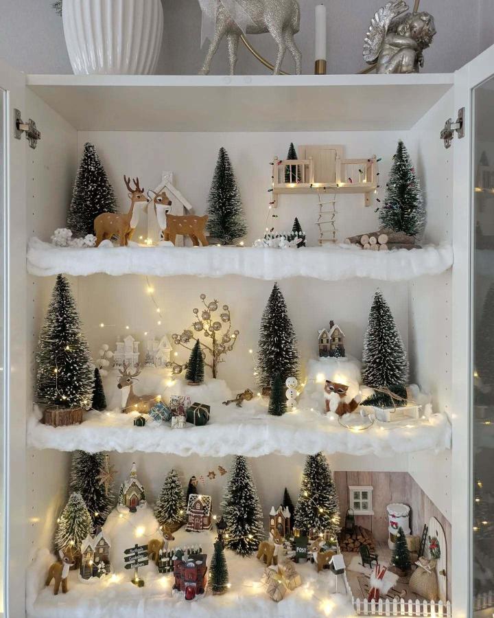 White tiered shelving display from @idylliskasagoland featuring bottle brush trees, deer figurines, and snowy village scenes with fairy lights.