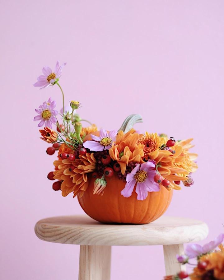 Orange pumpkin vase from @loreak.co filled with dahlias, cosmos, and berry stems on wooden stool.