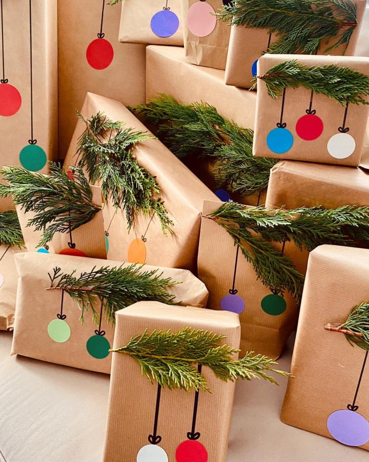 Kraft paper wrapped gifts from @hecho_en_casa_a_mano with hand-painted colorful ornament doodles and fresh greenery sprigs.