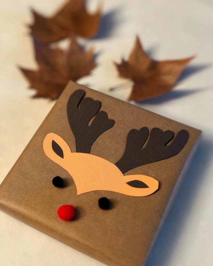Kraft paper gift with construction paper reindeer face from @hecho_en_casa_a_mano featuring pom-pom nose and antlers.