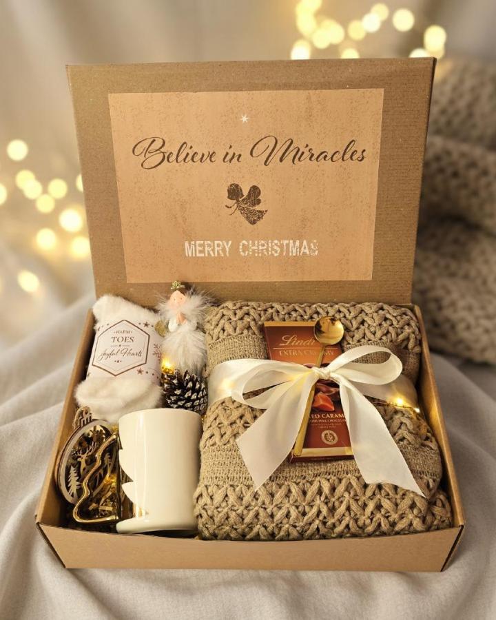 Kraft gift box from @giftingcy with cozy slippers, woven basket, hand cream, and festive ribbon.