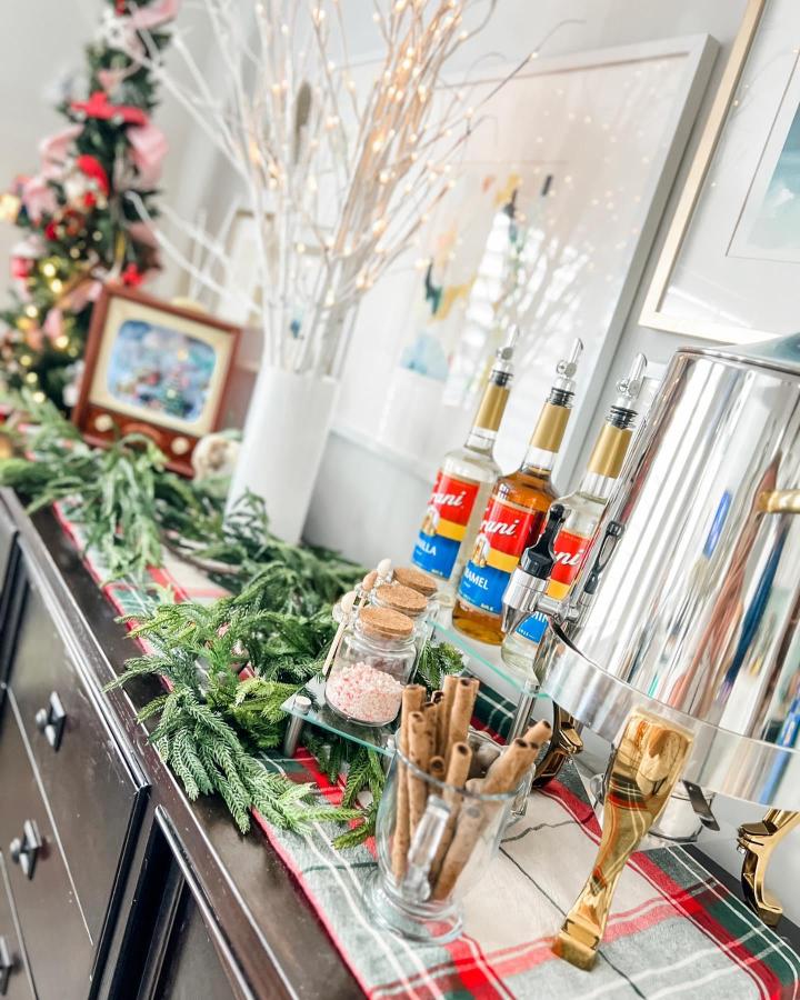 White lighted branches and plaid runner hot cocoa station from @eventsby.janelle with syrup dispensers and greenery.
