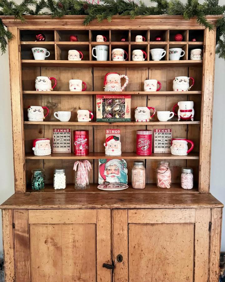 Rustic wooden hutch from @prettyandprintedshop displaying festive mugs, ornaments, and seasonal drink station decor