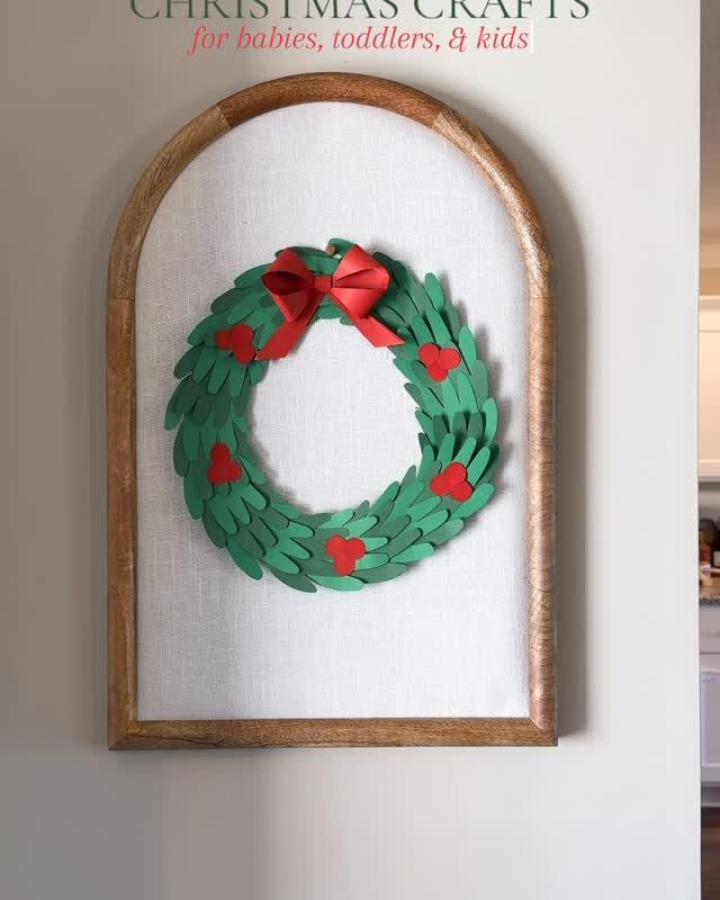 Green paper handprint wreath from @nat.hinson with red hearts and ribbon bow in arched frame.