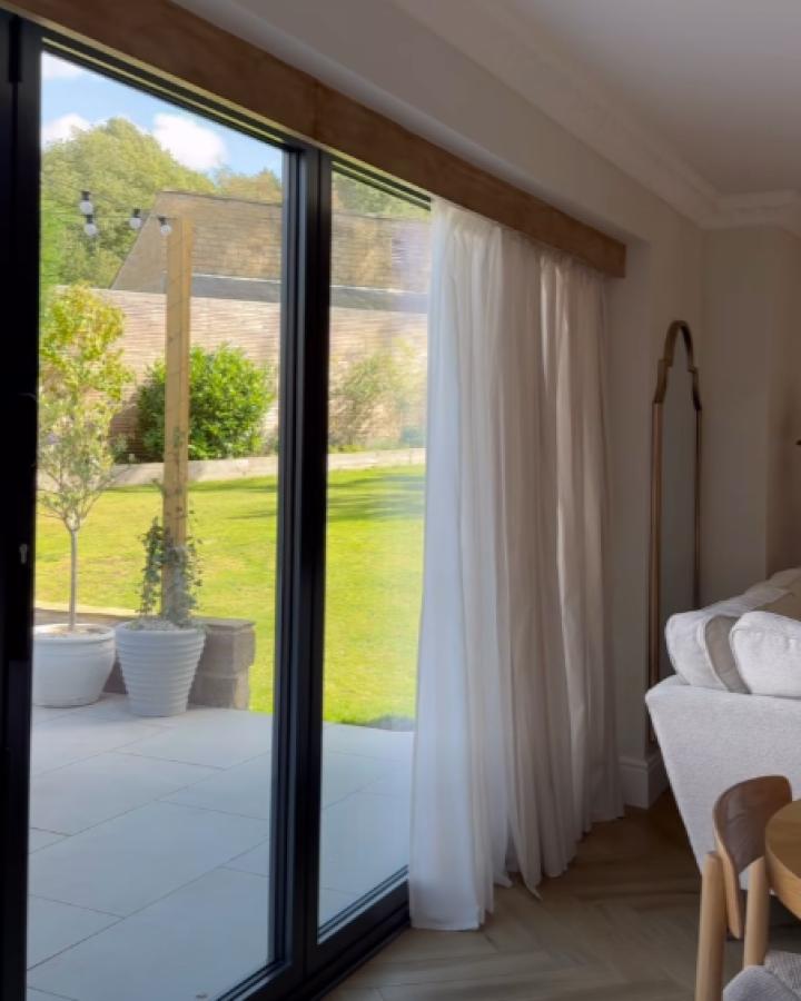 Wooden curtain pelmet from Klk.interiors above patio doors with white drapes and garden view.