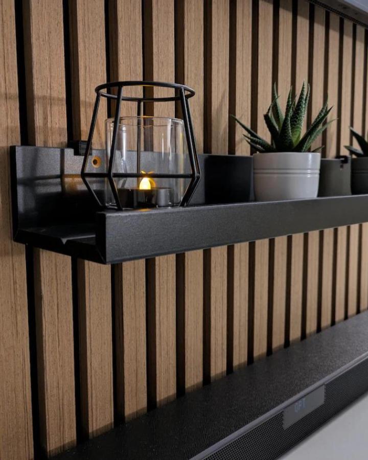 Black acoustic panel brackets from GroovyGiftCo holding Mosslanda shelf against textured wood wall with candles and plants.