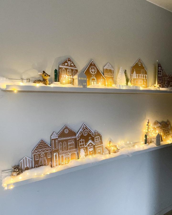 Illuminated cardboard gingerbread village display from @modernehjemsnapchat. Two shelves feature handmade paper houses with string lights.