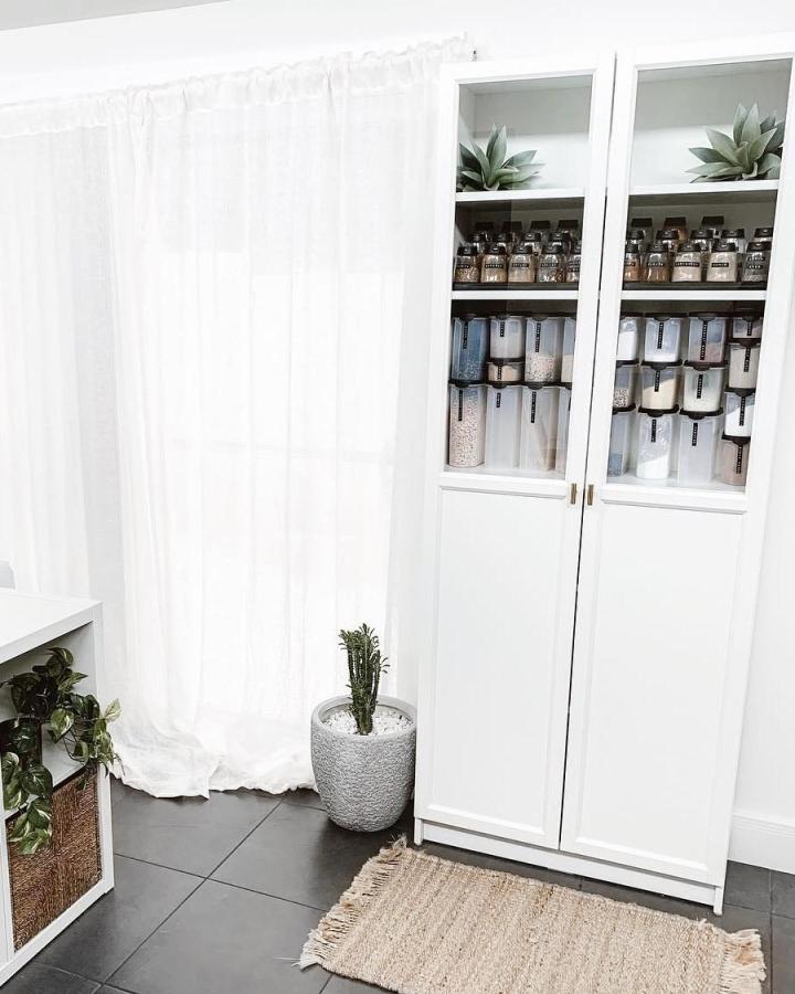White glass-front pantry cabinet from @_ashlee_white_. Labeled clear containers for ikea pantry ideas, with brass pulls and plants.