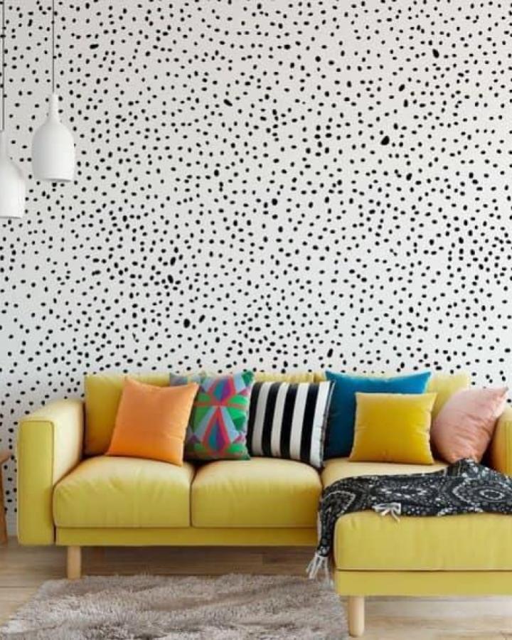 Black and white spotted peel & stick wallpaper ideas on living room wall behind yellow couch with colorful pillows.