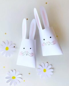 white paper bunny cones with pink ears and hand-drawn faces surrounded by paper daisies and gold stars easter paper crafts for adults from @luciamallea