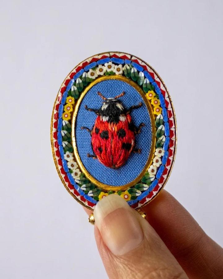 Bright embroidered ladybug brooch with floral border on blue fabric background from @megembroiders showcasing ladybug crafts for adults.