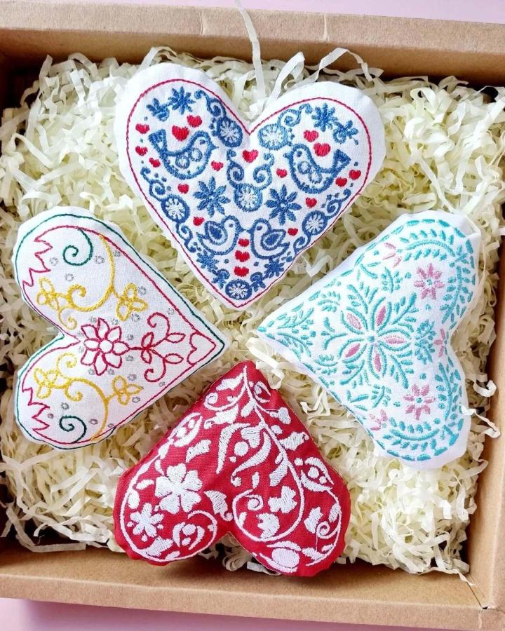 Four embroidered fabric hearts with floral and bird motifs nestled in shredded paper gift box valentines crafts for adults from @emelshandicraft. Hand stitched with colorful thread on cotton.