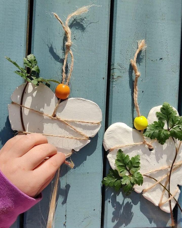 white clay hearts wrapped with twine and adorned with green leaves and colorful beads valentines crafts for kids from @mudnbloom hanging on a blue wooden surface