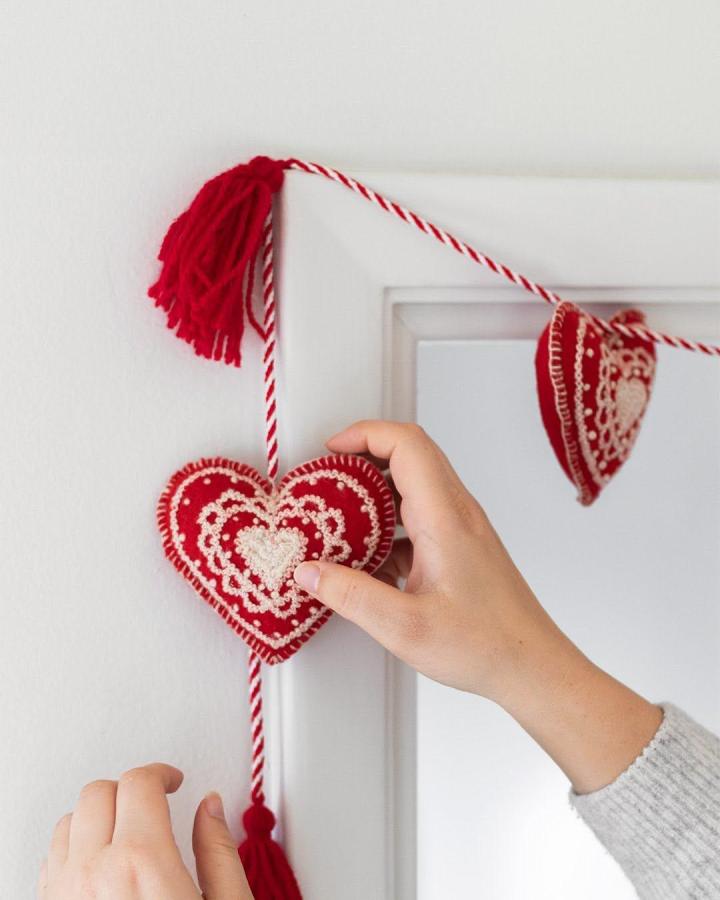 Red felt hearts with cream embroidery hanging on twisted red and white cord garland from @heirloomartco valentines heart garland ideas with yarn tassels