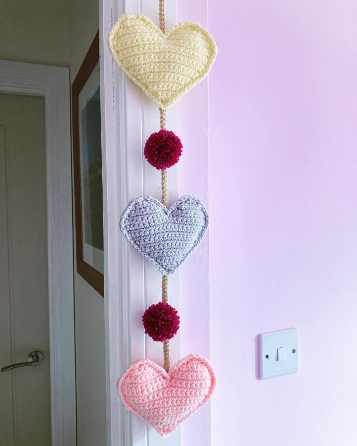Crocheted pastel hearts and raspberry pom poms garland from @coffee_and_crochet_24 hanging by a doorway for valentines heart garland ideas.