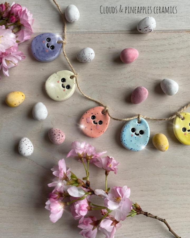Pastel speckled clay eggs garland with smiling faces from @pottdpeople on wooden surface with pink flowers easter clay craft ideas.