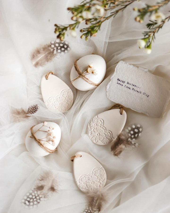 Off-white handmade clay Easter egg tags with lace texture and natural twine from @amberstextiles on soft tulle background with feathers.