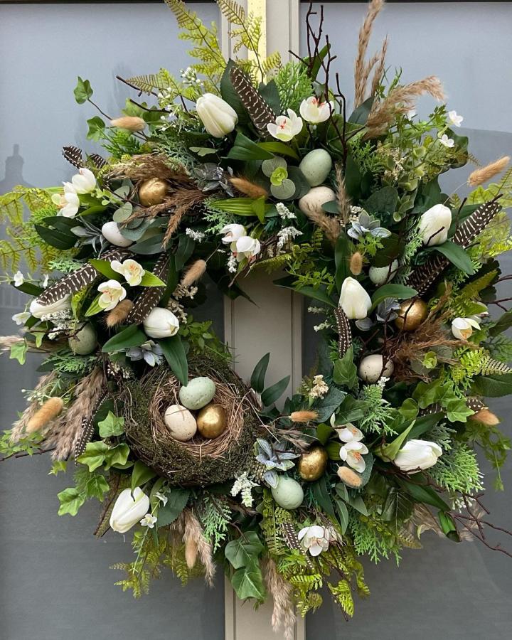 Green leafy Easter wreath with white tulips, feathers, and a bird nest decorated with pastel and gold eggs from @alanas_floral_designs.