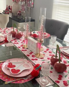 Glossy red ceramic cherry centerpieces and playful cherry print table runner from @our.cozy.little.home valentines table decor ideas with pink candles and glassware.