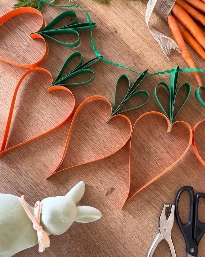 Orange and green quilled paper carrot hearts Easter garland ideas from @nowherenearmartha displayed on a wooden table with crafting tools.