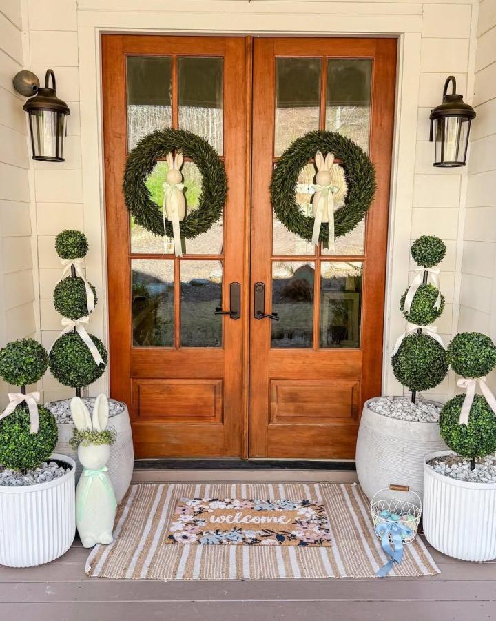 Green boxwood wreaths with bunny figures and topiary pots on a wooden porch from @montgofarmhouse Easter porch decor ideas with natural tones and ribbons.