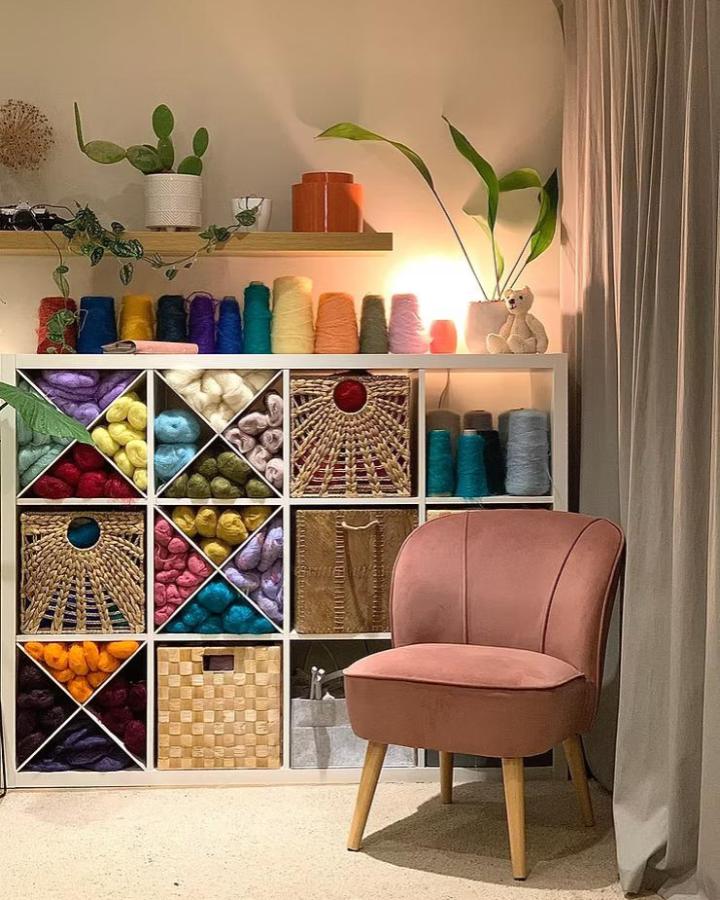 Soft pink chair beside white ikea kallax craft room ideas yarn organizer with diagonal dividers and woven baskets from Knittedly. Cozy and colorful craft space.