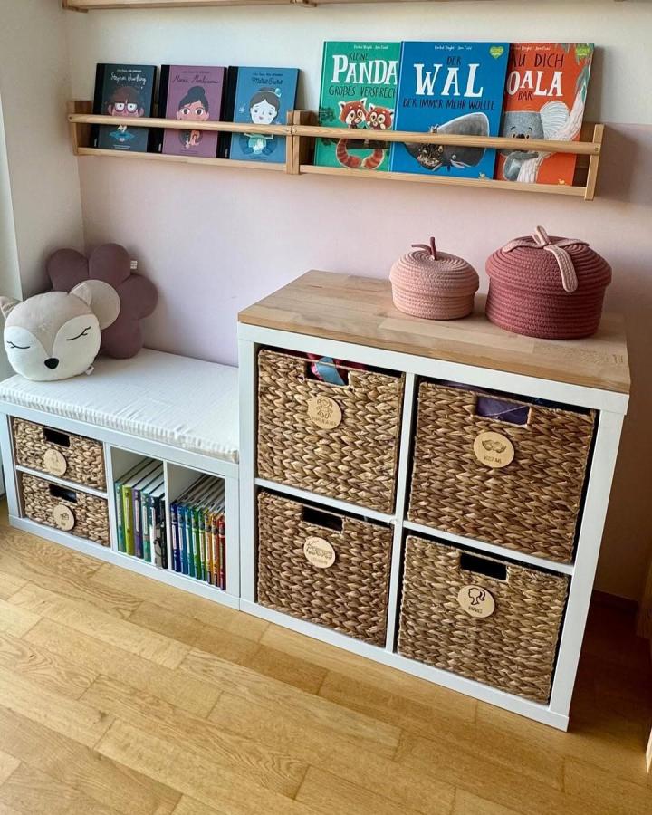 woven white and wood toy storage from @ordnung_kinderleicht. Kallax cubbies with wicker baskets, books, and bench cushion.