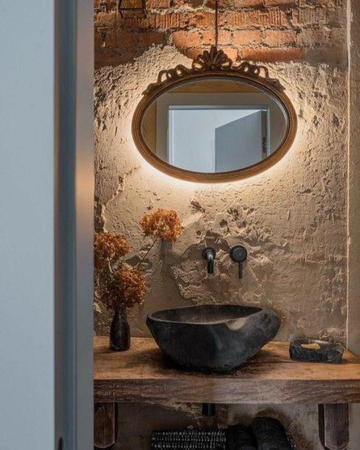 Vintage gold oval mirror with subtle backlighting in rustic small bathroom by IVD.ru. Rough plaster wall and black stone sink complement each other.