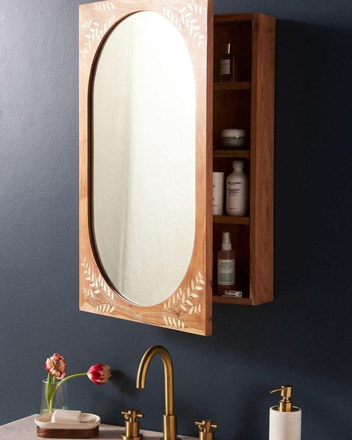 Wood-framed oval mirror with leaf carvings and hidden shelving from Anthropologie for bathroom mirror ideas for small spaces. Mounted on dark wall above gold faucet.