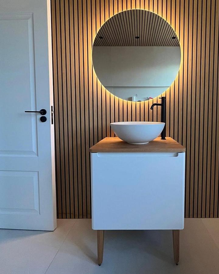 Vertical wood slat bathroom wall panel ideas from @neutralsandnoire with round mirror and minimalist vanity lighting.