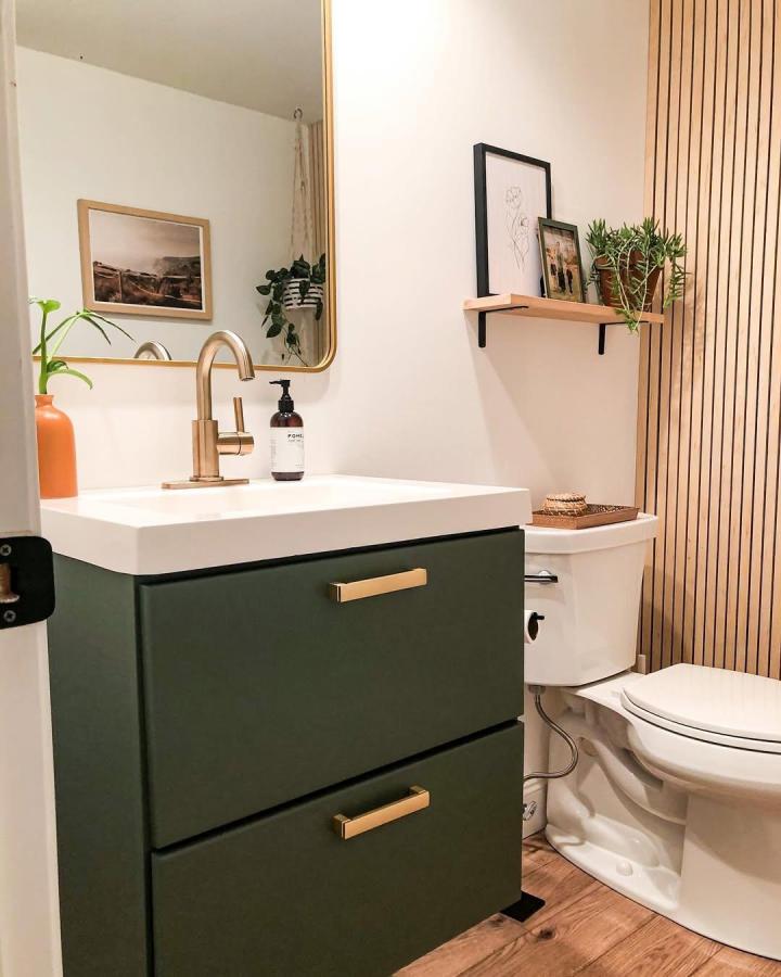 Vertical natural wood slat bathroom wall panel ideas from @urbanoaksdesignco with green vanity and brass fixtures create warm inviting space