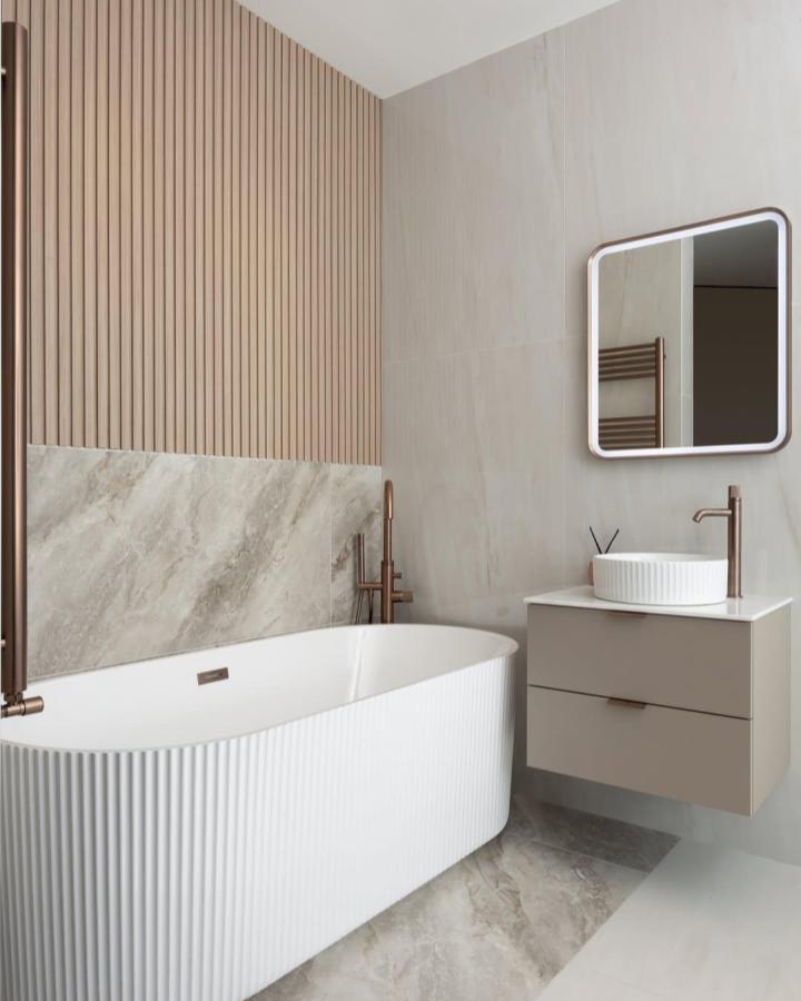 Neutral wood slat panels and marble-look tiles in a bathroom from @tilemerchant with bronze fixtures and soft beige tones.