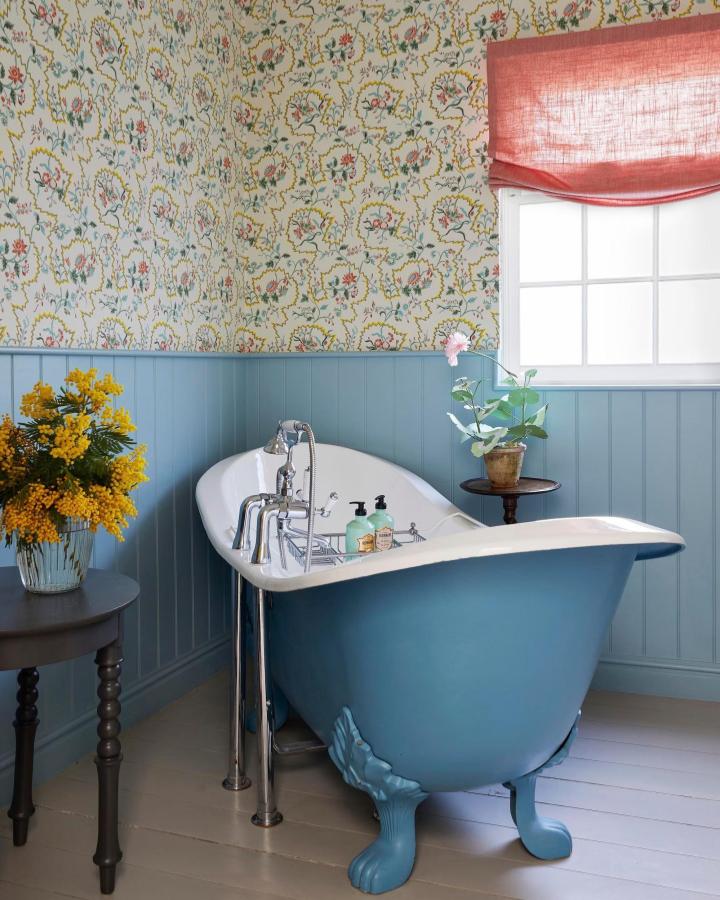 Blue painted clawfoot tub and paneling in charming bathroom from Country Living with floral wallpaper and natural light.