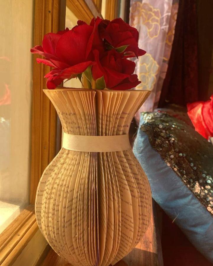 Vintage folded book pages shaped into a vase holding bright red flowers for classic book page home decor crafts. Natural light highlights paper texture and flower color.
